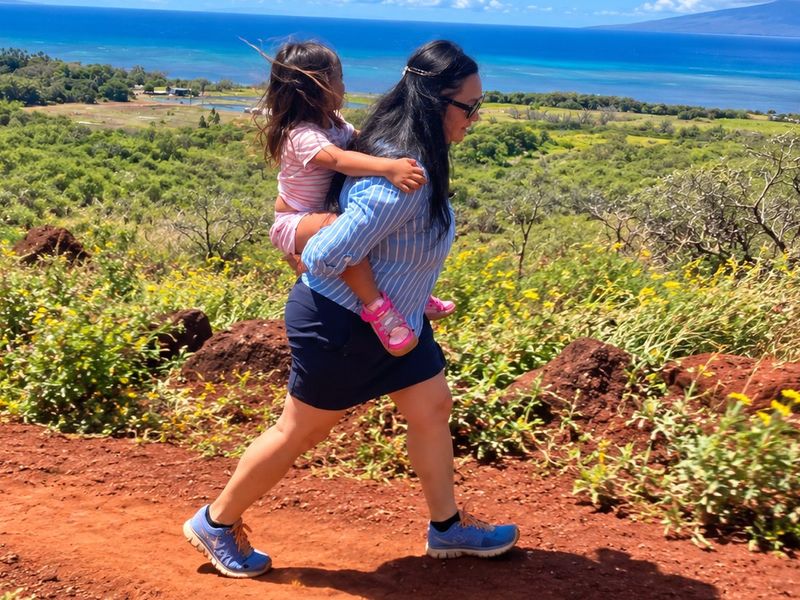 A woman carrying a child on her back along a red-dirt trail above the coast