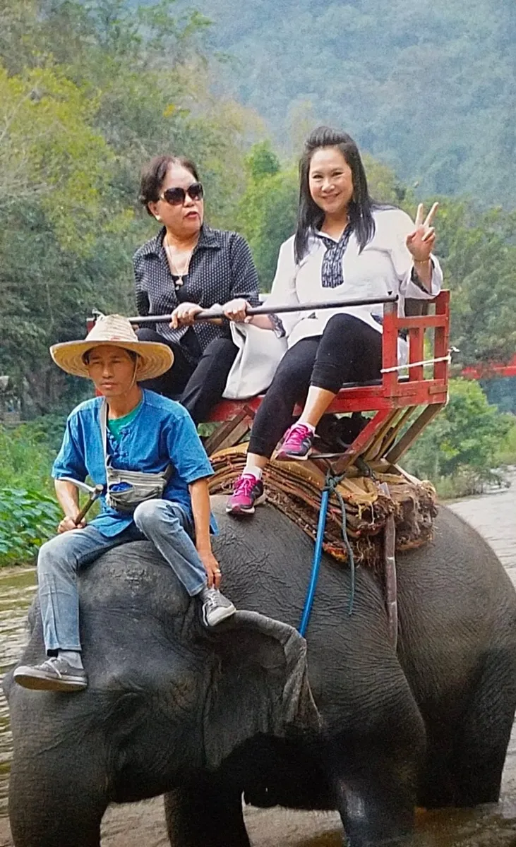 Two women and a guide riding an elephant across a river, with forested hills behind