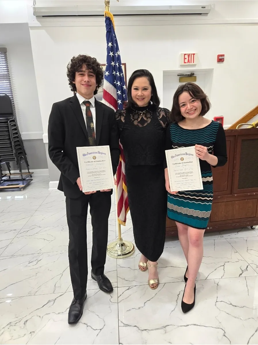A woman stands between two young people holding certificates, beside an American flag
